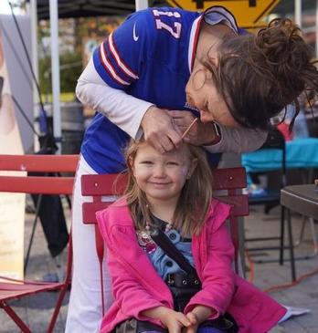 A smiling young girl in a pink jacket sits on a red chair as a woman, dressed in a blue sports jersey, painting her face.