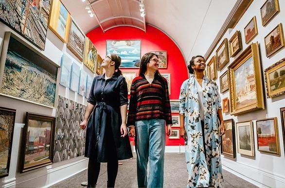 Three people walking through an exhibition in Museum London, Ontario observing many paintings hung on the walls.