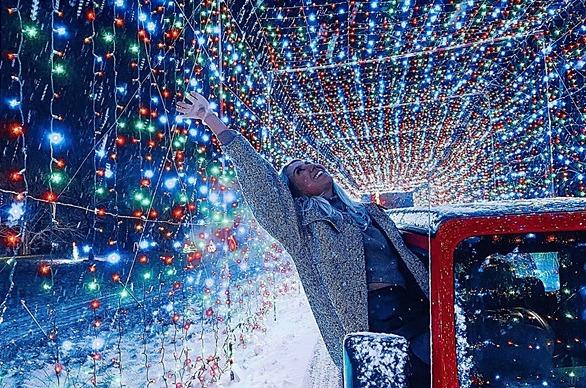 A female stretching out from the passenger side of a vehicle while its driving through a tunnel of coloured Christmas lights