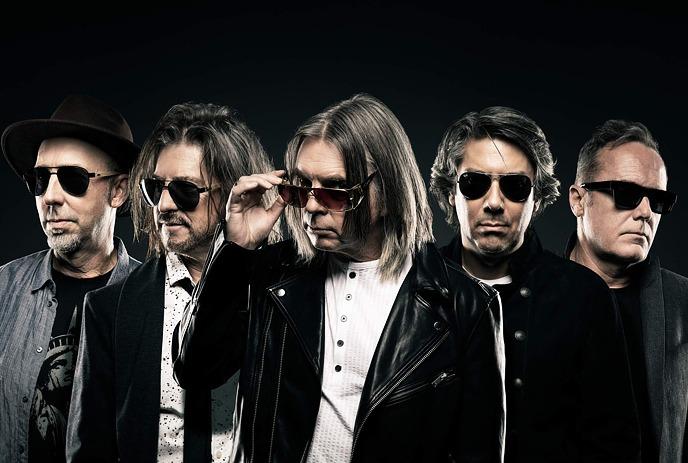 A group of five men wearing sunglasses, dressed in jackets, stand together in rock band style pose against dark background.