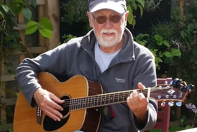 A person is sitting outdoors playing an acoustic guitar, with an accordion on a nearby chair.