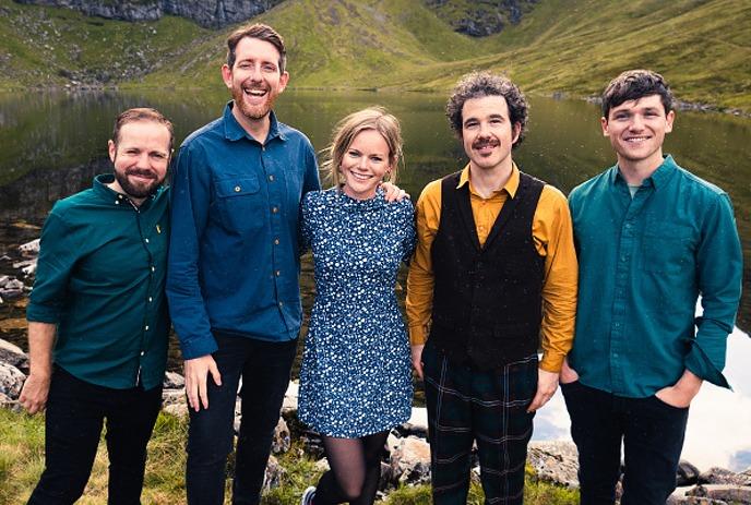 A group of five smiling people stands outdoors, dressed in colorful clothing, with a scenic lake and hills in the background