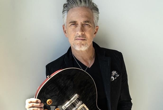 Colin James, in a black jacket, leaning against a wall while holding a black and red Gibson electric guitar.