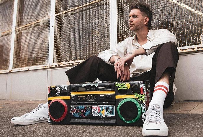 A person sitting on the ground with a colorful boombox covered in stickers, wearing white sneakers and striped socks.