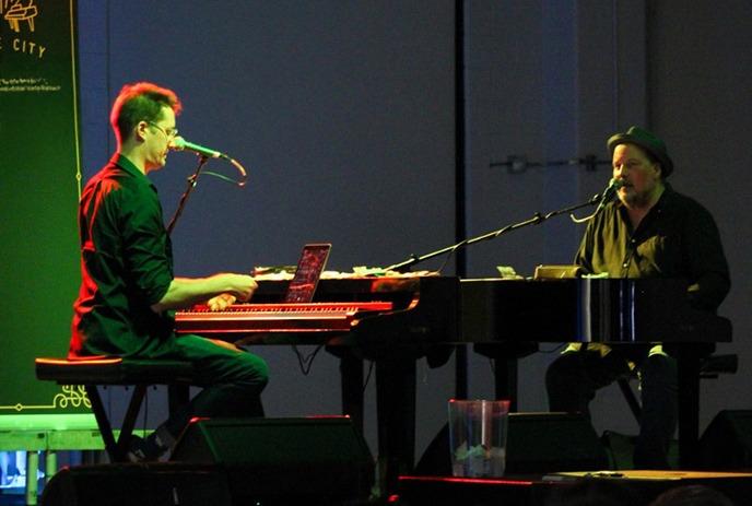 Two musicians are performing on stage with pianos under green spotlights.