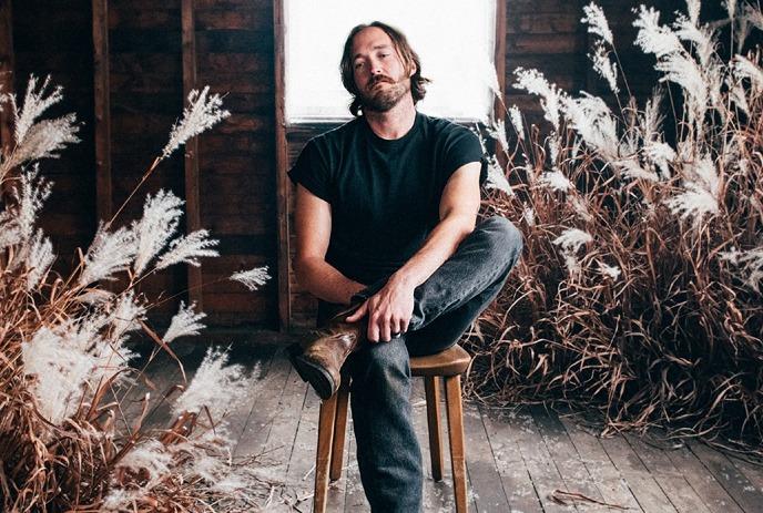 An artist on a wooden stool in a rustic room with wooden floors and walls, decorated with dried plants and a large window.