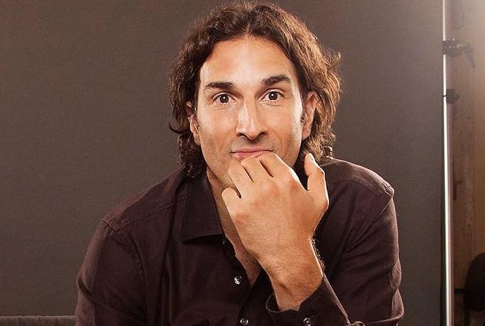 Comedian Gary Gulman sitting down posing for the camera, in front of a brown background.
