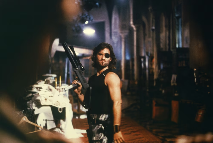 A man with an eyepatch, holding a shotgun, stands confidently in a dimly lit, industrial setting with dark background.