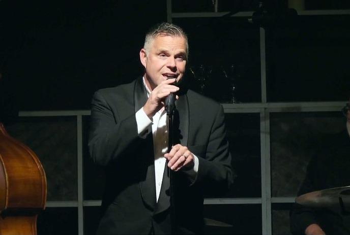 A person in a tuxedo is speaking into a microphone, with a blurred face. The background features a grey curtain.