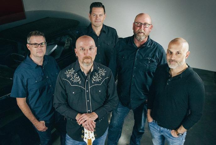 A group of five men, all in dark shirts, pose together for a photo. Two of them are bald, and one holds a guitar.