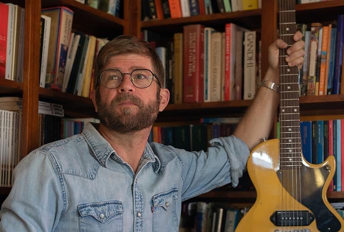 A man in a denim shirt with a yellow guitar posing for the picture with books at the background.