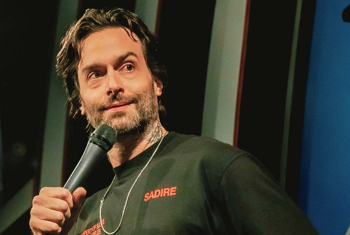 Chris D'Elia wearing a green shirt, holding a microphone on stage.