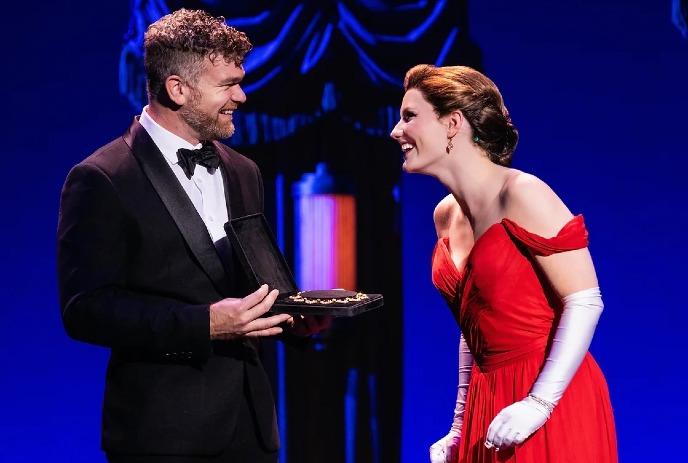 A man and a woman wearing formal attire, the man holding open a jewelry box, and the woman smiling at him.