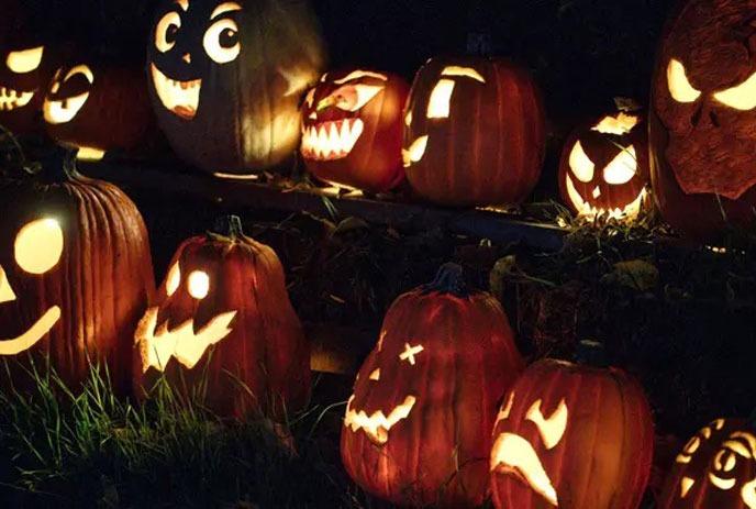 A group of people in Halloween costumes gather in a dimly lit room, enjoying drinks, with a carved pumpkin centerpiece.