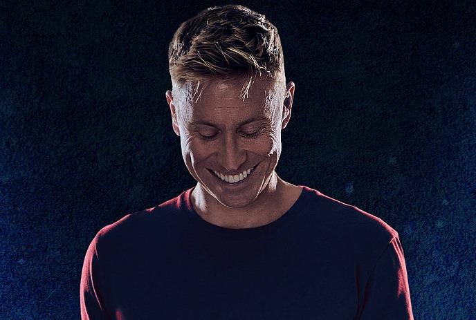 Russell Howard looking at the ground smiling, in front of a blue background.