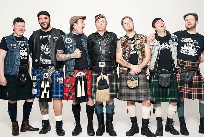 The 7 band members of The Real McKenzies standing against a white wall, posing for the camera, wearing kilts.