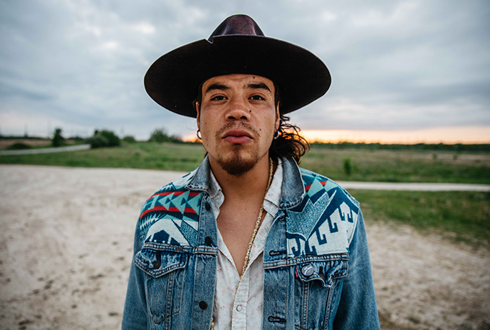 Logan Staats standing in a field wearing  jean jacket and a hat.