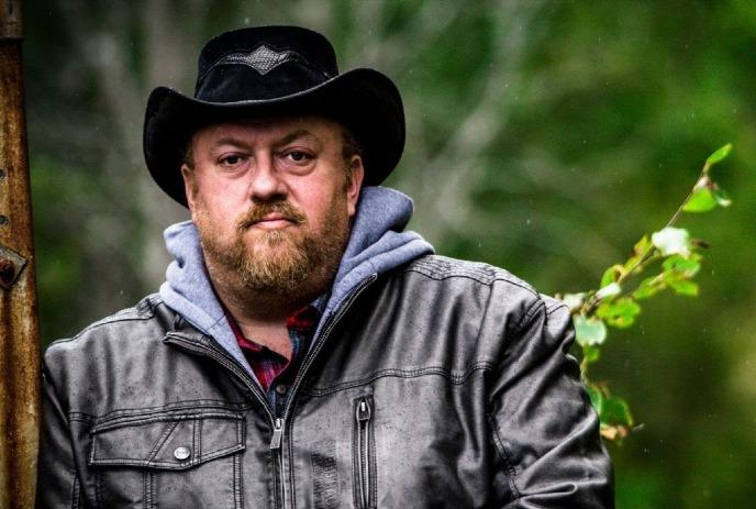 J.P. Cormier wearing a black hat and a leather jacket, under a green background.