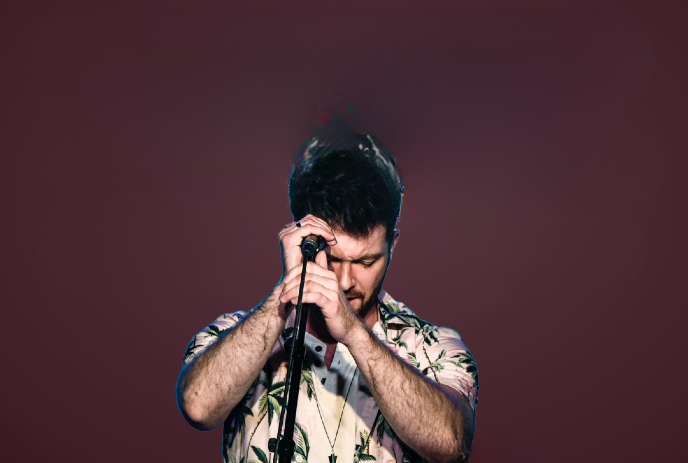 A man, with closed eyes, singing passionately into a microphone. He is wearing a floral shirt, with a dark background.