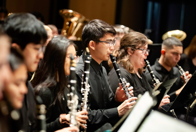 A group of musicians in formal attire playing wind instruments like clarinets and brass, with a dark background.