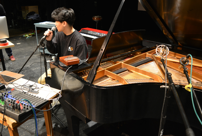 A person singing into a microphone beside a grand piano with a laptop and audio equipment on a table.