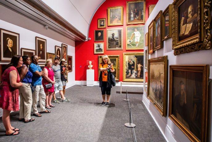 A group of people attentively listening to a tour guide in an art gallery, surrounded by portraits and historical paintings.