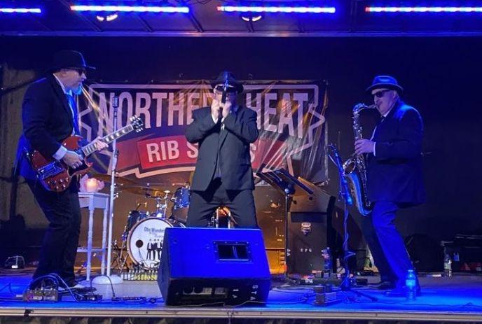 Band performs on stage at night, lit by blue lights, with “Northern Heat Rib Series” banner in the background.