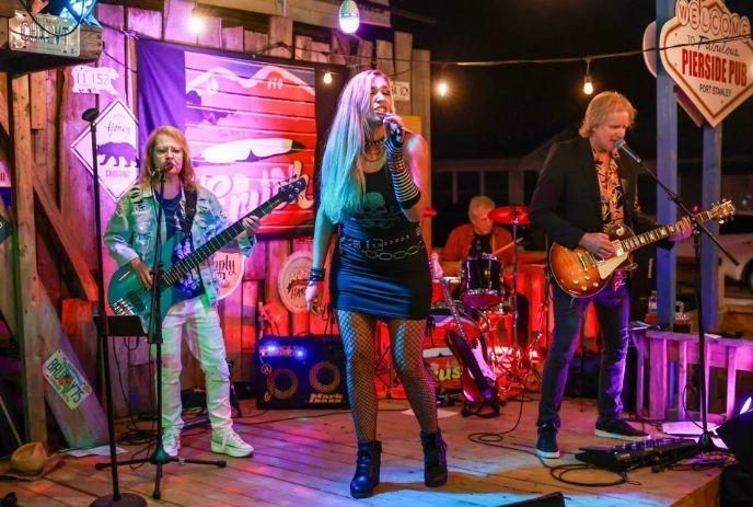 A band performing on a wooden stage. The female vocalist wears black, and the bassist, guitarist, and drummer.