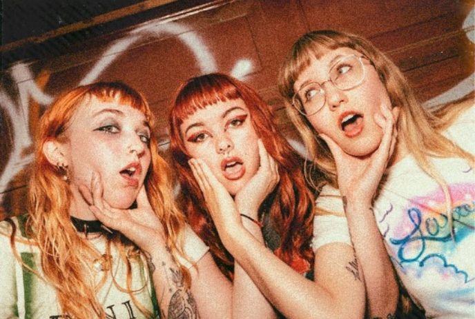 Three young women with red and blonde hair are posing closely together. Each of them is holding their faces.