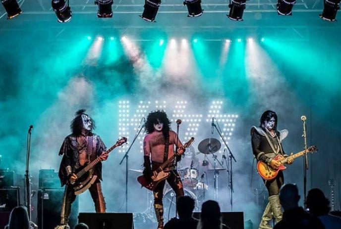 Musicians dressed as iconic rock band KISS perform on stage, with vibrant blue and green lights and a foggy atmosphere.