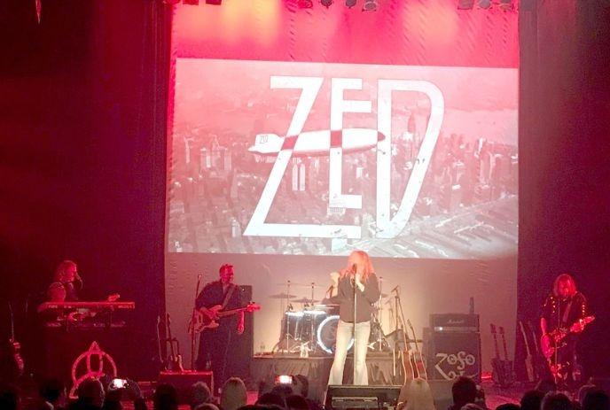 A live performance by the band Zed, featuring a vibrant stage setup with red lighting, a backdrop displaying their logo.