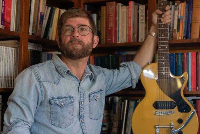 A man in a denim shirt with a yellow guitar posing for the picture with books at the background.