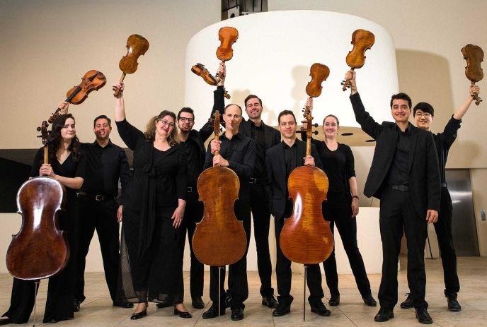 A group of musicians in black attire holding up string instruments, including violins and cellos, in a celebratory pose.