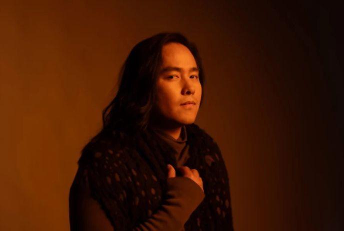 A person with a warm-toned background, wearing a dark, textured shawl and a long-sleeved shirt posing for the picture.