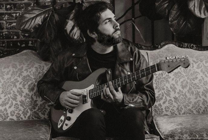 Artist in a leather jacket is sitting on a patterned sofa, playing an electric guitar. The face is blurred. The background includes large leaves and a brick wall.