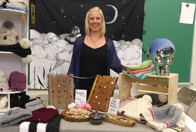 A Woman stands behind a table of handmade crafts, including knitted hats and jewelry, with a moon tapestry backdrop.