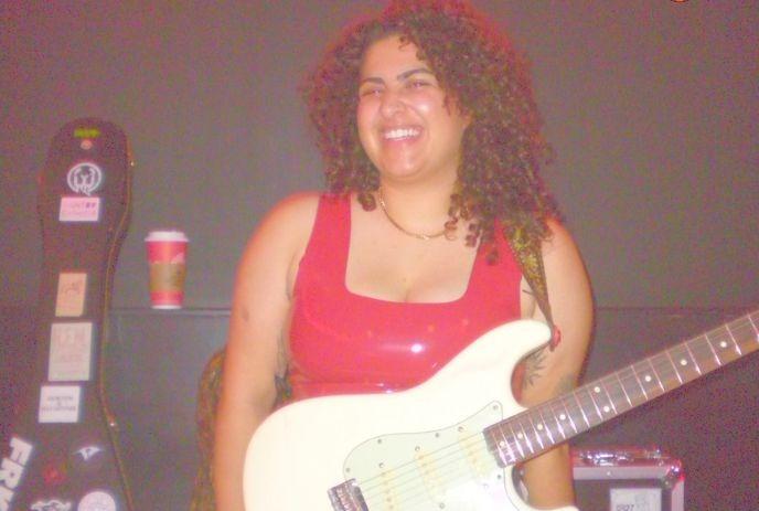 Lady artist in a red top holds a white electric guitar; background includes a guitar case and coffee cup.