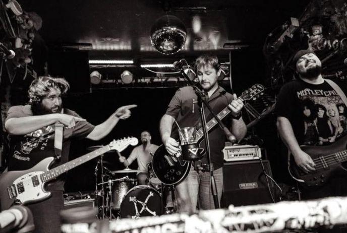 Live band on stage in black and white: drummer, guitarist, bassist performing amidst bright lights and musical equipment.