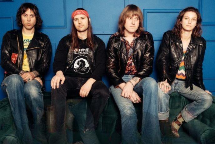 Four band members in leather jackets sitting on a couch under a blue wall posing for a picture.