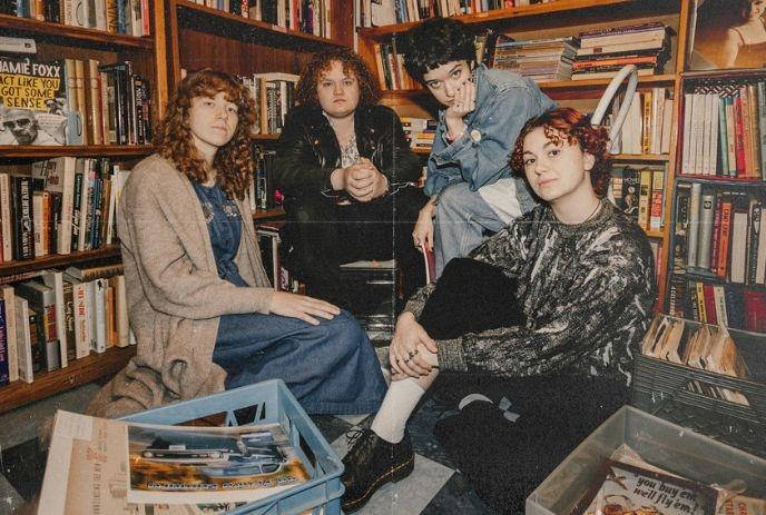 Four artists are seated on the floor, surrounded by bookshelves, posing for a picture within a cozy setting.