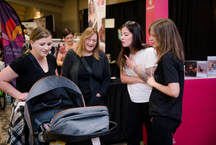 Four women at an indoor event gather near a stroller and booth, engaging in conversation and interactions.