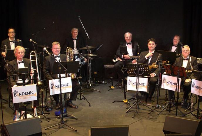 A band of musicians in formal attire with various instruments, including trombones, saxophones, drums, performing on stage.