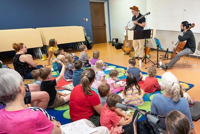 Two musicians playing to a group of children with their parents or care givers in a room