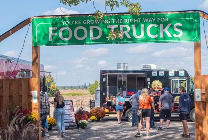 Outdoor food court with people at picnic tables, food trucks in the background, and string lights overhead.