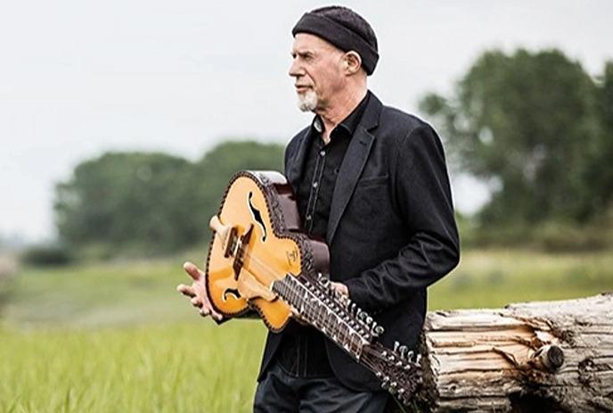 A person in a black suit playing a guitar while sitting on a log in a grassy field, with trees and green in the background.