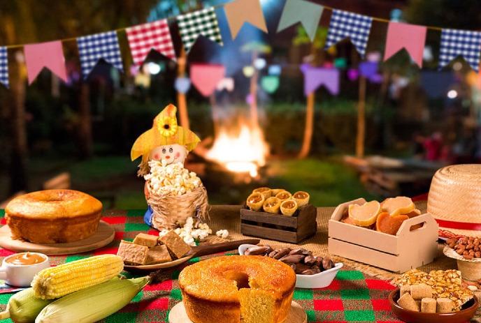A festive outdoor setting at night. An array of food items, including corn on the cob, cakes, pastries, cookies, and nuts.