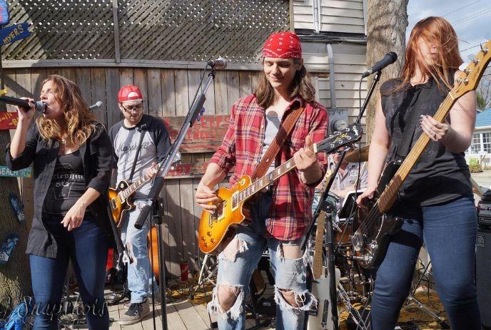 A band performs outdoors with a singer, two guitarists, and a bassist on a wooden stage with a wooden fence backdrop.