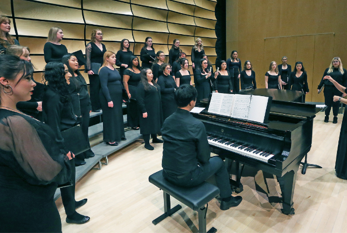 A choir dressed in black performs on risers with a pianist playing a grand piano, directed by a conductor in a concert hall.