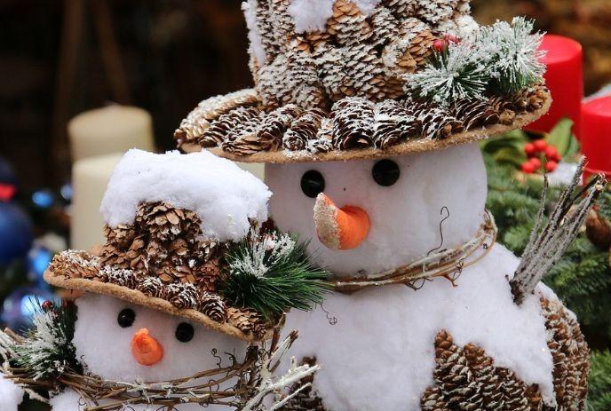 Two snowmen made of pinecones and snow, wearing hats, with carrot noses & twig arms, surrounded by festive decorations.