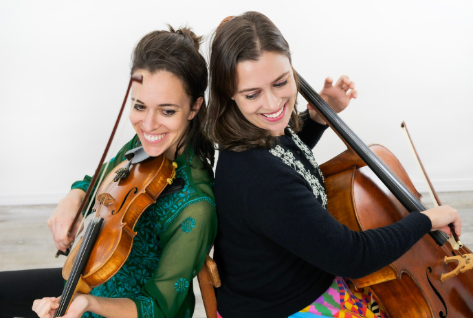 Two musicians with smiling faces sitting side by side playing a violin and a cello under a white background.